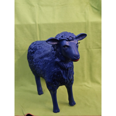 Das blaue Schaf Stehende Schafskulptur mit leuchtend blauem, strukturiertem Fell, dunklen Beinen und einem schmalen Tiergesicht vor grünem Hintergrund.