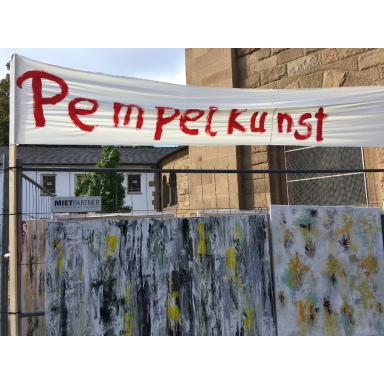 Kunst unter freiem Himmel Banner mit der roten Aufschrift Pempelkunst über mehreren abstrakt bemalten Tafeln bei einer Ausstellung im Freien.