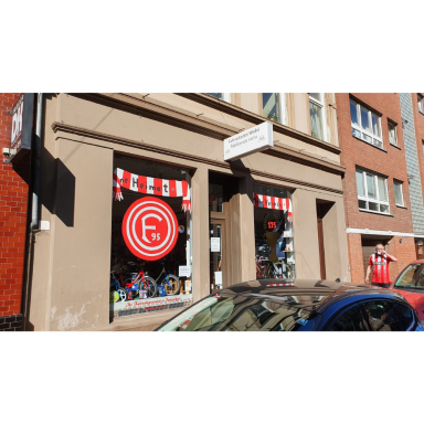 Kunst im Schaufenster I Schaufenster eines Gebäudes mit rot-weißer Markise und einem großen rot-weißen runden Emblem; im unteren Bereich sind gestaltete Objekte zu
