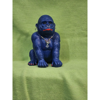 Der blaue Gorilla Sitzende dunkelblaue Gorillaskulptur mit kräftigen Armen, roten Lippen, roten Zehennägeln und goldfarbener Halskette vor grünem Hintergrund.
