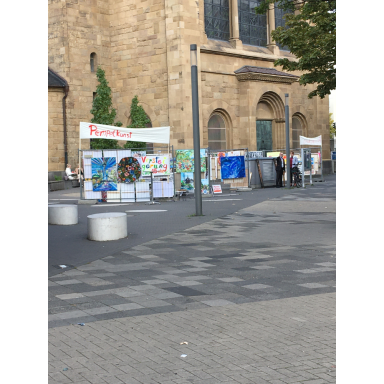 Kunst auf dem Platz Auf einem gepflasterten Platz vor einem historischen Steingebäude sind mehrere farbenfrohe Bilder und Ausstellungstafeln aufgestellt.
